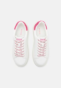 Baskets blanches avec des accents roses sur le talon, présentant une tige texturée, des lacets blancs avec un branding 'GUESS' répété, et une toe arrondie.