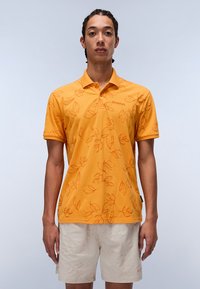 Gul polo trøje med bladmønster i en lysere nuance, korte ærmer og krave. Båret med lyse shorts.