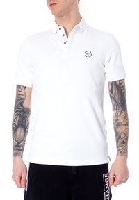 Hvit polo-skjorte laget av bomull med en tre-knappers placket og brodert logo på brystet. Korte ermer med ribbede mansjetter.