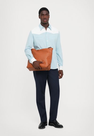 Camicia blu e bianca con colletto a punta, tiene una borsa in pelle marrone. Pantaloni scuri e scarpe nere completano l'outfit.