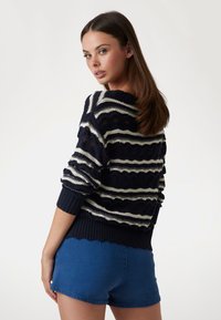 Maglione lavorato a maglia blu navy con strisce ondulate bianche e un motivo testurizzato, dotato di polsini a coste e orlo ondulato, abbinato a pantaloni corti in denim.