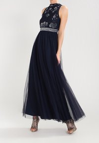 Granatowa długa sukienka z wysokim kołnierzem, haftowaną górą i zwiewnym tiulowym dołem. Posiada srebrne akcenty i przezroczystą nakładkę.