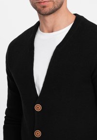 Černý pletený kardigan s výstřihem do V, který má tři hnědé knoflíky a texturovanou látku. Nositelný přes bílé tričko.