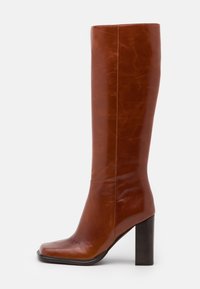 Botte en cuir marron jusqu'au genou avec un bout carré, un haut talon bloc et une texture lisse. Présente des détails de couture sur les côtés.
