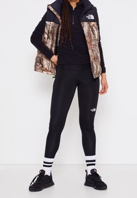 Leggings neri, top nero e un gilet a motivo camouflage con colletto alto. Sneakers nere con calze a righe bianche.