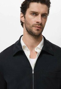 Black zip-up jacket with a collar, front pocket, and smooth texture, layered over a white shirt, with a plain background.