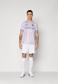 Homme se tenant face, portant un maillot de football violet clair avec "vonovia" et un logo, un short blanc, des chaussettes, des crampons et des tatouages visibles sur les bras et les jambes.