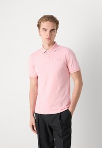 Mladý muž s krátkými vlasy, který má na sobě světle růžové polo tričko s krátkým rukávem a černé kalhoty, stojí s jednou rukou v kapse.