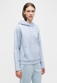 Γυναίκα που φοράει γαλάζιο φούτερ με κουκούλα και παντελόνι που ταιριάζει, με λευκό λογότυπο Emporio Armani EA7 στα μανίκια και στο πόδι του παντελονιού.