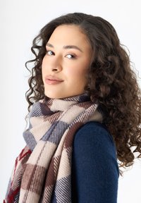 Écharpe à carreaux en beige, marron et bleu marine. Tissu doux drapé avec style, offrant un look superposé. Le mannequin porte un pull bleu marine et a des cheveux bouclés.