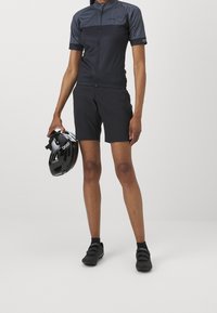 Kvinnors cykeloutfit i svart, med en kortärmad zip-up topp och matchande shorts. Håller en svart hjälm med vita accenter.