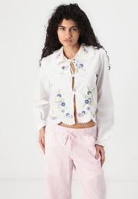 Weiße Bluse mit floralem Stickerei, gewelltem Saum, Bindeschluss und langen Ärmeln. Kombiniert mit strukturierten rosa Pyjamahosen.