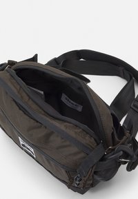 Brun nylon budväska med ett dragkedjad huvudfack, justerbar rem och extern etikett. Har en texturerad tyg och minimal hårdvara.