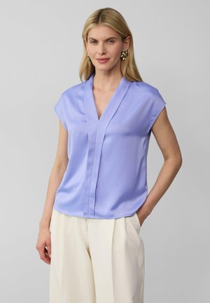 Blouse violette clair à manches courtes avec un décolleté en V, texture lisse et détails discrets de couture à l'avant, associée à un pantalon couleur crème.