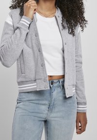 Person wearing light blue jeans, white crop top, and gray varsity jacket with white stripes, holding the jacket collar with one hand.