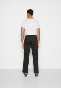 Black straight-leg pants with back pockets, worn with a white t-shirt. The model is wearing green sneakers, seen from the back.