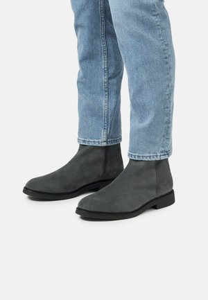 Botas de tobillo de ante gris con un diseño minimalista, puntera redonda, suela plana y detalles de costura sutiles, combinadas con jeans de mezclilla azul claro.