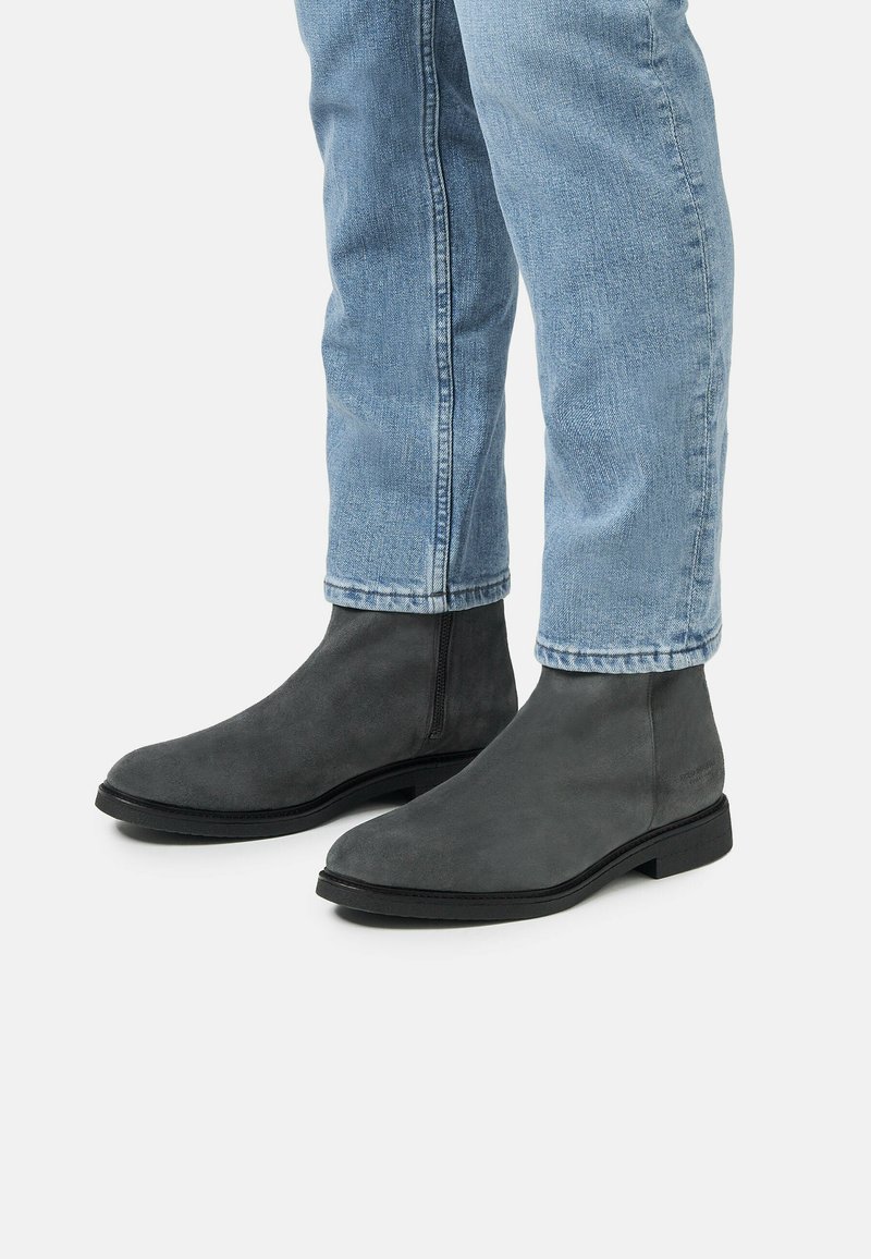 Bottines en suede gris au design minimaliste, à bout rond, semelle plate et détails de couture discrets, portées avec un jean bleu clair.