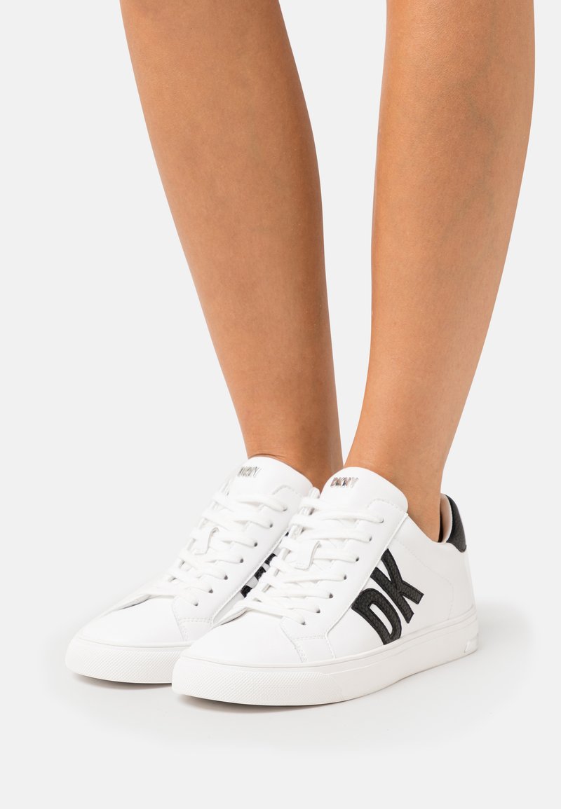 Hvite sneakers med svart DK-logo, snøring foran, teksturert svart hæltab, gummisåle, glatt lær overdel og minimalistisk design.