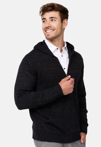 Schwarze Strick-Zip-Hoodie mit geräumiger Kapuze, gerippten Bündchen und Fronttaschen, über einem weißen Hemd mit Knöpfen getragen.