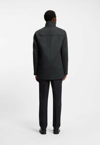 Man in een donkergrijze wollen jas, zwarte dress pants en zwarte schoenen, staand met zijn rug naar de camera tegen een effen witte achtergrond.