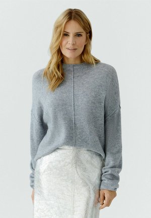 Blonde vrouw met een losse grijze trui ingestopt in een metalen zilveren rok, staand tegen een effen witte achtergrond.