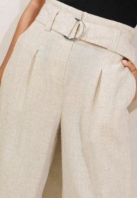 Pantalones de lino beige con un diseño de pierna ancha, que presentan pliegues frontales y una gran hebilla circular plateada en un cinturón a juego.