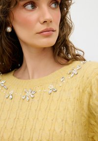 Maglione giallo lavorato a maglia con un motivo testurizzato, dotato di accenti decorativi in cristallo e perle attorno al collo.