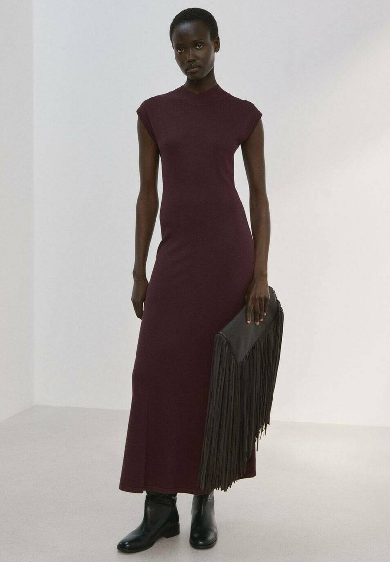 Femme portant une robe longue sans manches bordeaux, bottines noires, tenant une grande pochette noire à franges dans une pièce blanche et vide.