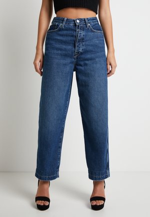 Donna che indossa jeans blu a vita alta e gamba larga con tasche frontali e chiusura con bottoni, abbinati a tacchi neri aperti e top nero corto.