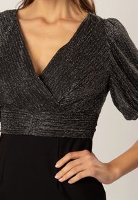 Femme portant une robe noire avec un corsage ajusté pailleté, des manches courtes bouffantes et un décolleté en V profond, la main reposant sur la hanche.