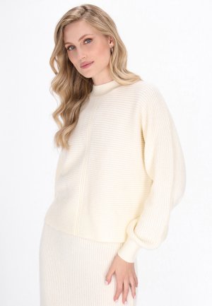 Crème-kleurige gebreide trui met een hoge kraag, geribbelde textuur en ballonsleeves. Bevat subtiele verticale naden en heeft een cropped snit.