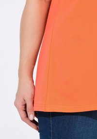 Camiseta de manga corta naranja hecha de algodón suave, con dobladillo recto y un sutil detalle en la costura lateral, combinada con jeans azul oscuro.