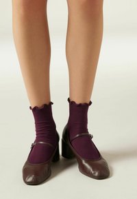 Brązowe skórzane buty typu Mary Jane z paskiem i klamrą, połączone z prążkowanymi purpurowymi skarpetkami o falbaniastych krawędziach.