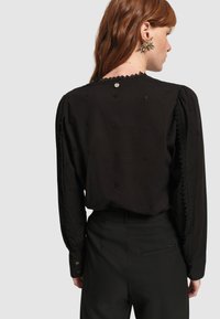 Blusa nera con dettagli floreali ricamati, accentuata da volant lungo il colletto e i polsini, maniche lunghe e chiusura a bottone lucido sulla schiena.