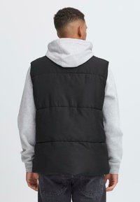 Schwarze gepolsterte Weste mit hohem Kragen, getragen über einem grauen Hoodie. Sie hat eine gesteppte Textur und ist für zusätzliche Wärme in Schichten konzipiert.