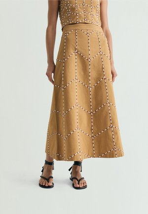 Mujer con falda color tostado con patrones geométricos blancos y marrones cosidos, combinada con sandalias planas negras con tiras.