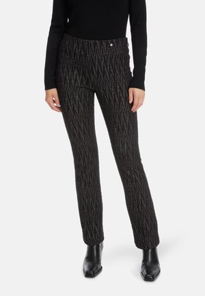 Schwarze gemusterte Hose mit strukturiertem Finish, die ein Zickzackdesign und zwei vordere Knöpfe aufweist. Kombiniert mit schwarzen Knöchelstiefeln.