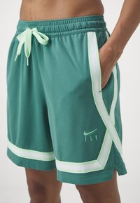 Person trägt türkisfarbene Sportshorts mit weißen und hellgrünen Streifen, Kordelzugbund und "Nike Fly"-Logo nahe der linken Tasche.