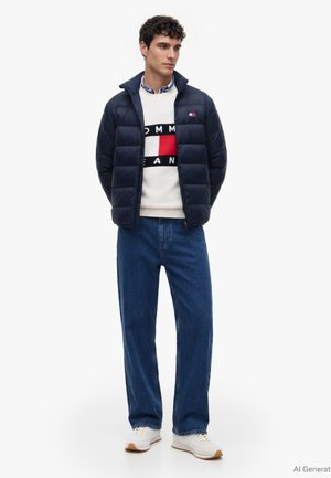 Man draagt een navy pufferjack, witte sweater met rood en zwart logo, blauwe spijkerbroek en witte sneakers, staat met handen achter rug.