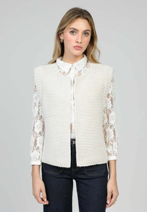 Jeune femme portant un gilet blanc sans manches en tricot par-dessus un chemisier blanc en dentelle florale et un jean foncé, debout devant un fond uni.