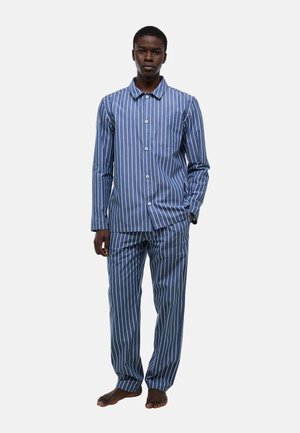 Jeune homme debout pieds nus portant une chemise à manches longues boutonnée à rayures verticales bleues et blanches et un ensemble de pyjama assorti.