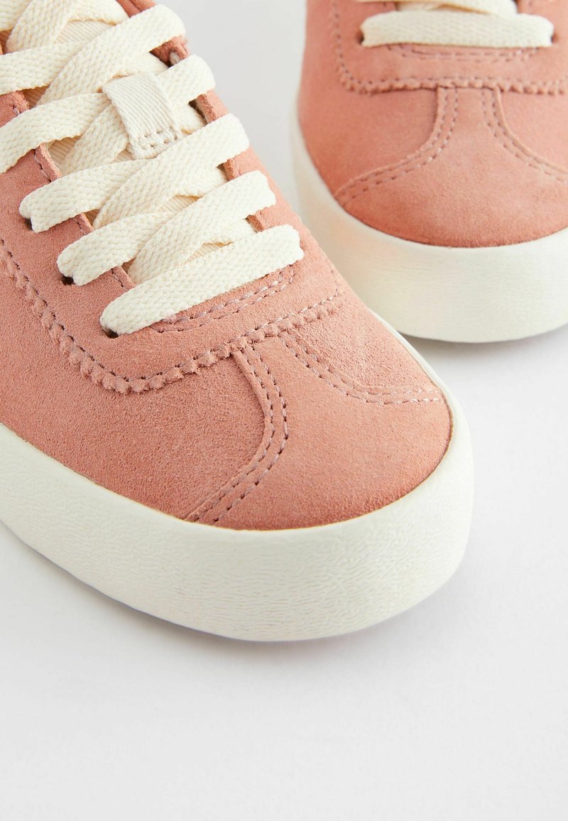 Zapatillas de ante rosa con cordones crema, puntera redonda, suela de goma blanca y costuras decorativas en la parte superior. Vista en primer plano de los detalles del diseño.