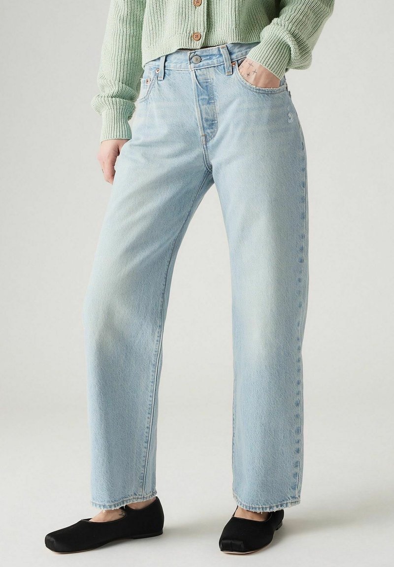 Personne portant un jean droit bleu clair, un cardigan vert boutonné, des chaussures plates noires, debout avec une main dans la poche.