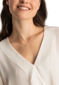 Goldene Kette mit einem herzförmigen Anhänger, der ein strukturiertes Design aufweist, getragen von einer Person mit einer sanften, hellfarbigen Bluse.