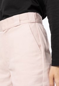 Světle růžové kalhoty s vysokým pasem, bočními kapsami a viditelnou texturou látky. Stylizované s černým topem s dlouhým rukávem, který je částečně vidět.