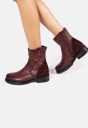 Bottes basses en cuir bordeaux avec une fermeture éclair latérale, un bout rond et des accents de panneaux texturés, ornées d'un bouton décoratif en laiton.