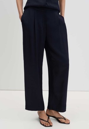 Person trägt lockere dunkelblaue Hose mit Händen in den Taschen und schwarze Flip-Flop-Sandalen mit Absatz und steht auf einem hellen Boden.