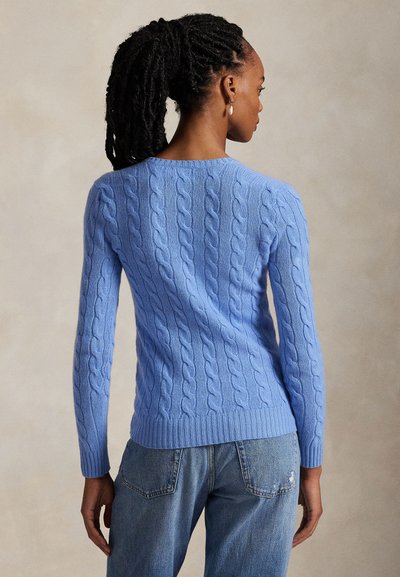 Sötét fonott haját hátul összekötő nő világoskék kötött mintás pulóvert és kék farmert visel, háttal egy semleges háttér előtt.