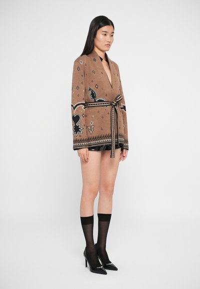Cardigan marron à motifs avec un décolleté en V profond, taille ceinturée et accents noirs. Porté avec des talons hauts noirs et des chaussettes transparentes montantes.
